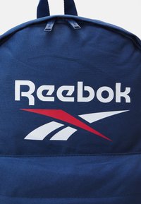 Sötétkék hátizsák tartós szövetből, amelyen kiemelkedő fehér Reebok logó és piros részletek találhatók egy letisztult dizájnon. Két cipzárhúzó a tetején.
