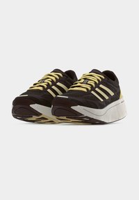 Tumepruunide spordijalatsite paar, millel on paksud valged tald ja kollased paelad, kollaste ja valgete triipude rõhutustega.