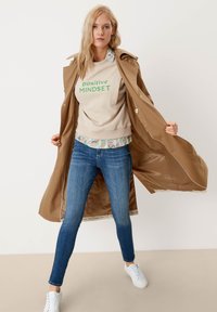 Ruskea trench-coat beige-hupparin päällä, jossa lukee "positiivinen AJATTELUTAPA", siniset farkut ja valkoiset tennarit. Kerroksellinen kukkakuvioitu paita hupparin alla.