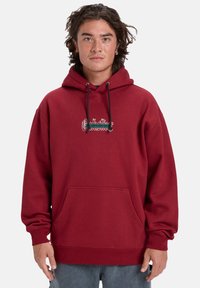 Maroon hoodie with a front pocket, drawstring hood, embroidered logo on the chest, and ribbed cuffs and hem. Cotton blend material.