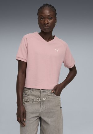 Mujer con una camiseta rosa claro de manga corta de Puma y jeans grises con un cinturón de cadena plateado en forma de estrella, sobre un fondo de gradiente gris.