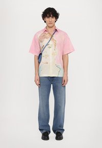 Camisa de manga corta rosa con un diseño de cara pintada, combinada con jeans de color azul claro y zapatos negros. Incluye un pequeño bolso cruzado azul.