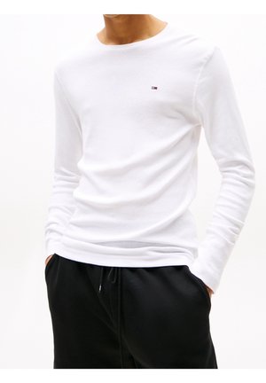 Camiseta blanca de manga larga con cuello redondo, confeccionada en tela suave. Cuenta con un pequeño logo bordado en rojo, blanco y azul en el pecho.