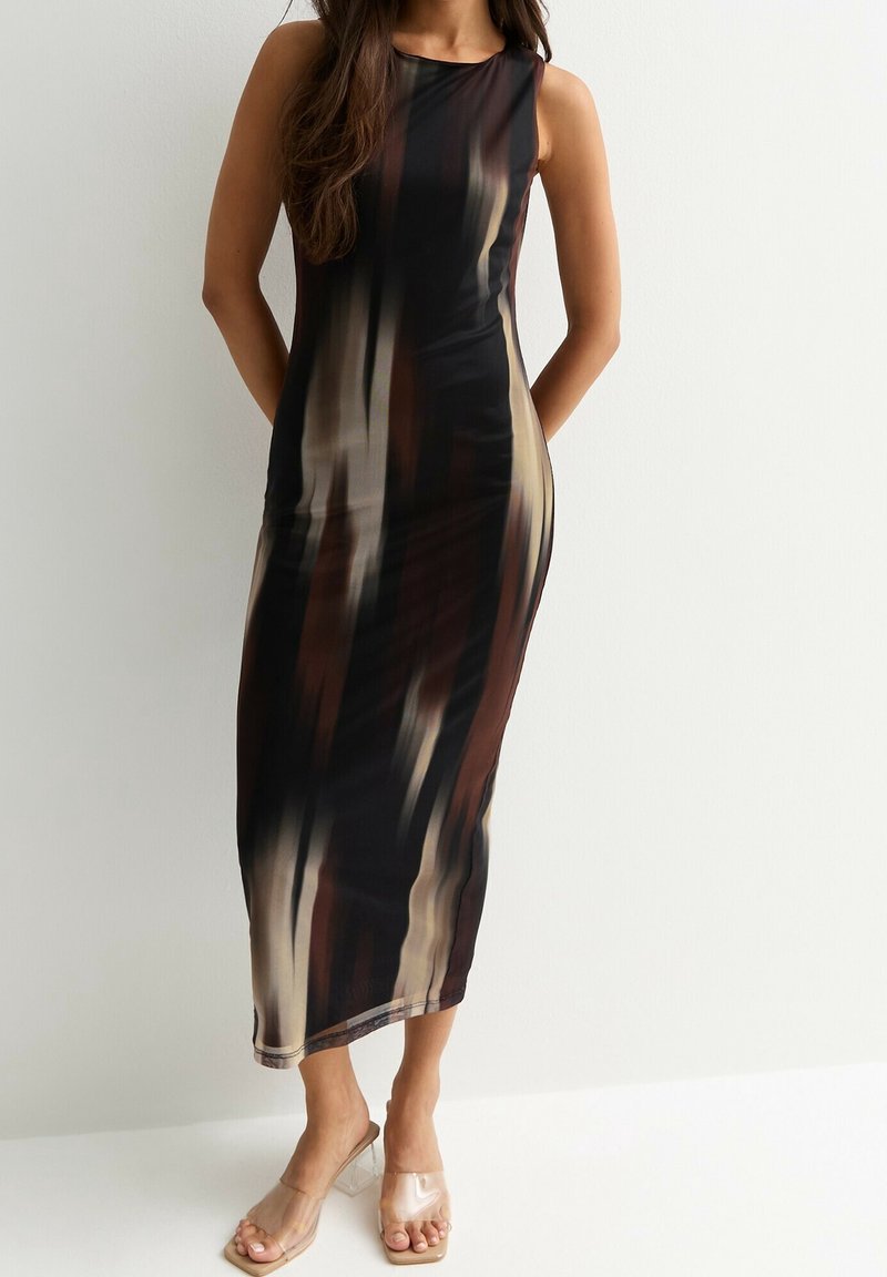 Femme en robe sans manches, longueur cheville, avec des rayures verticales marron, noires et beiges, portant des chaussures ouvertes à talons transparents, debout, mains dans le dos.