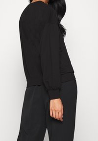 Haut noir à manches longues avec une coupe décontractée, des poignets côtelés et un col rond. Le tissu présente une texture lisse et un design minimaliste.