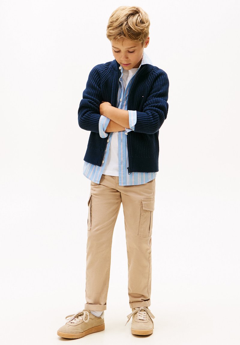 Marineblå strikket cardigan over en lyseblå stripete skjorte, khaki cargo bukser og beige sneakers. Enkelt design med ribbestruktur.