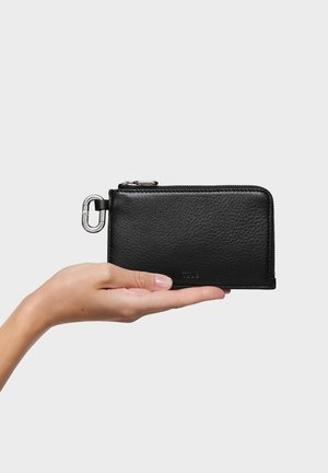 Portafoglio nero in pelle tenuto in mano, con chiusura a cerniera, ferramenta argentata e superficie testurizzata. Forma rettangolare compatta.