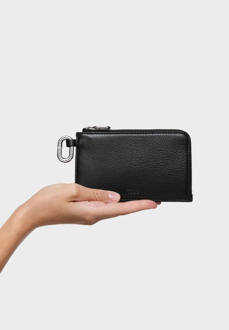 Czarny skórzany portfel trzymany w dłoni, z zamkiem błyskawicznym, srebrnymi elementami i teksturowaną powierzchnią. Kompaktowy, prostokątny kształt.