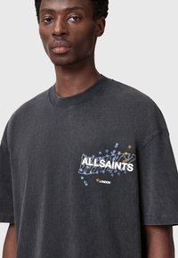 Sort oversized t-shirt i bomuld med et grafisk design, der indeholder teksten "ALLSAINTS" og farverige stjernesymboler på venstre bryst.