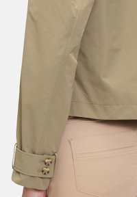 Licht olijfgroene jas met een gladde textuur, voorzien van knoopmanchetten en net gestikte naden, gecombineerd met pastelbeige broek.