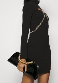 Femme dans une mini robe noire à découpes avec des détails en chaîne dorée, tenant une pochette noire avec une poignée en boucle dorée.