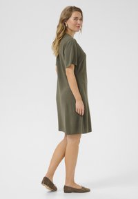 Robe olive verte à manches courtes, mi-longue, fabriquée en tissu doux avec une coupe décontractée. Associée à des chaussures plates marron, présentée de côté.