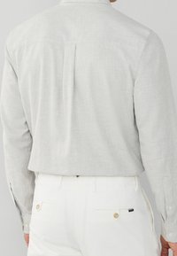 Camicia button-down grigia chiara a motivo, con maniche lunghe, infilata in pantaloni bianchi, che mostrano una texture liscia e un delicato motivo a quadretti.