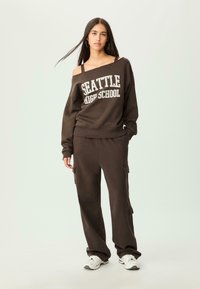 Felpa marrone con la scritta "SEATTLE HIGH SCHOOL", abbinata a pantaloni cargo coordinati. Il modello indossa sneaker bianche; tonalità terrose, tessuto casual.