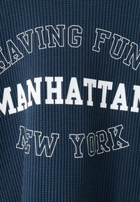 Temno modra teksturirana tkanina z belim napisom "IMAMO SE DOBRO MANHATTAN NEW YORK" v debelih in obrisanih pisavah.