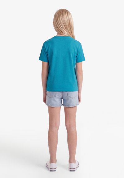 Παιδί με μακριά ξανθά μαλλιά που φοράει μπλουζάκι με κοντά μανίκια σε τυρκουάζ χρώμα, ανοιχτόχρωμα σορτς τζιν και λευκά παπούτσια τύπου slip-on, στέκεται γυρισμένο προς τα πίσω.