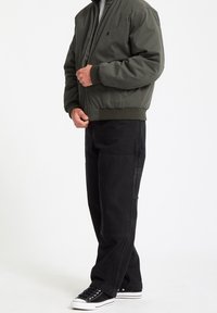 Personne portant une veste bomber vert olive, un pantalon cargo noir et des baskets basses noires, debout devant un fond blanc uni.