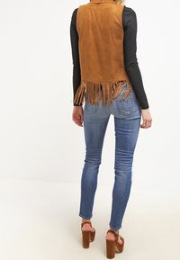 Chaleco de ante color cuero con flecos y corte ajustado, llevado sobre una camiseta negra de manga larga, combinado con jeans ajustados azules y tacones gruesos marrones.