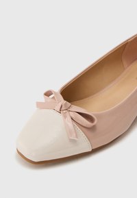 Ballet flat in blush pink and cream, featuring a glossy patent leather toe cap, a small bow detail, and a sleek, streamlined shape.