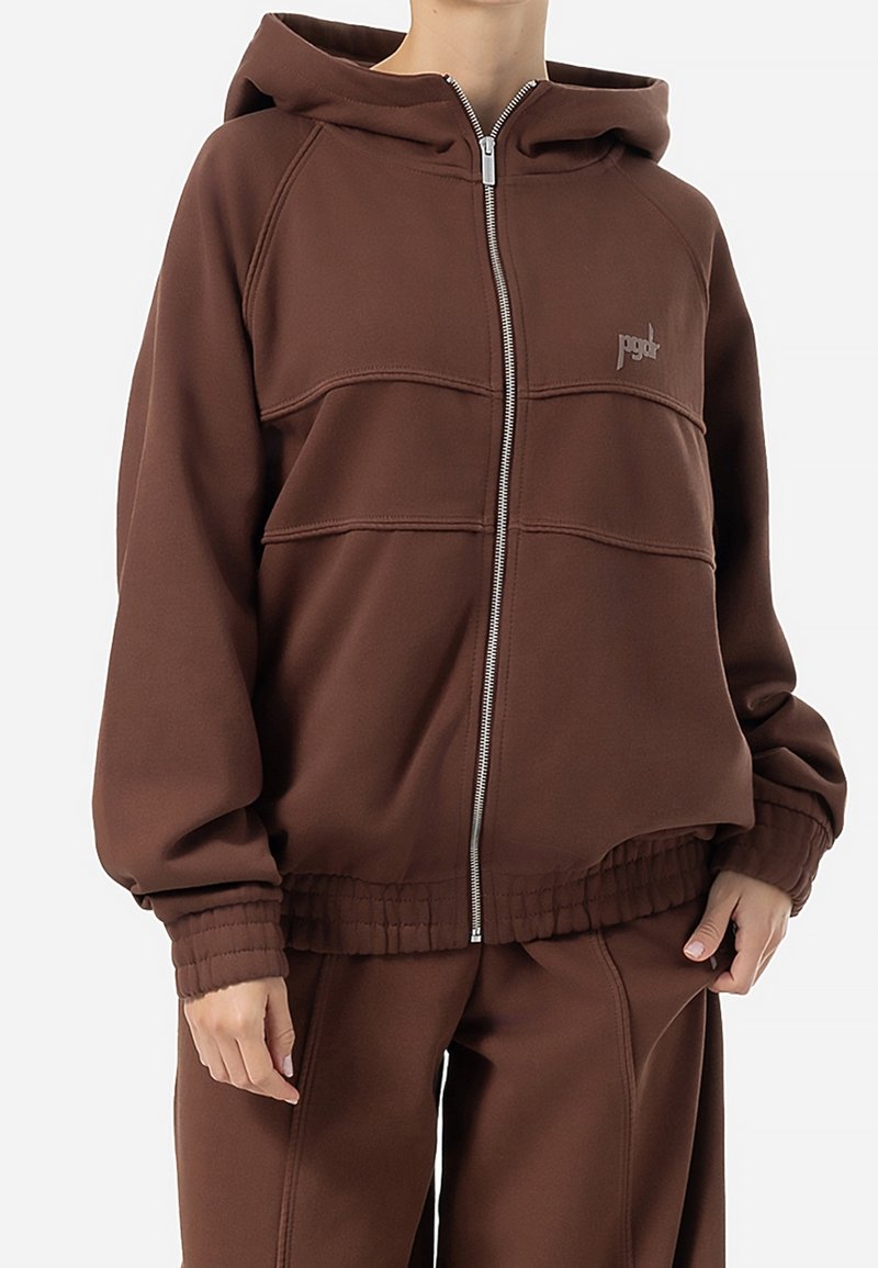 Sweat à capuche marron zippé avec capuche et taille et poignets élastiques, porté avec un pantalon marron assorti, avec un petit logo "pgdt" sur la poitrine.