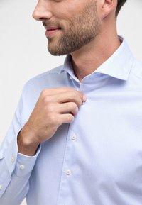 Camicia a righe blu chiaro con colletto a punta, polsini con un bottone e una texture di tessuto liscia. Vista ravvicinata della chiusura dei bottoni.