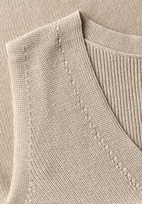Tessuto a maglia beige con una texture a coste, caratterizzato da cuciture piatte e un collo aperto e morbido. Il materiale sembra leggero e traspirante.