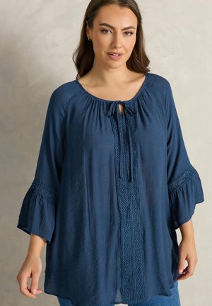 Kvinde iført en løs marineblå bluse med 3/4 klokkeærmer og blonde detaljer, stående foran en ensfarvet lys baggrund.