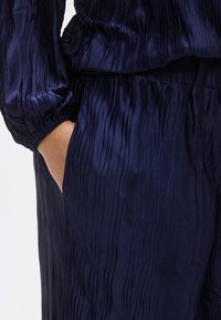 Tumesinise tekstuuriga kangast komplekt, mis sisaldab volditud särki pikkade varrukatega ja sobivaid pükse, millel on külgedel taskudetail.