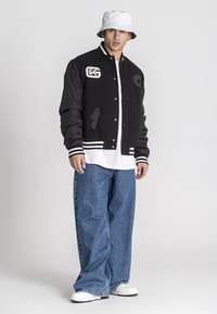Čierna varsity bunda s bielymi pruhmi a vyšitými nášivkami, skombinovaná s oversized modrými džínami a bielymi teniskami. Viditeľný klobúk bucket.