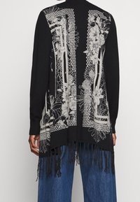 Svart kofta med intrikat vitt blommönster och geometrisk broderi, med fransdetaljer vid fållen och långa ärmar.