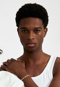 Lähedalt kujutatud inimene, kellel on lühikesed keerulised mustad juuksed ja valge top seljas. Kaadris on nähtaval käsivarrel kuldne käevõru.