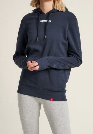 Personne portant un sweat à capuche bleu marine Hummel avec cordons et des leggings gris, les mains jointes devant, sur un fond uni.