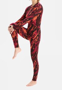 Pitkähihainen kokopuvun haalari, jossa on kirkas abstrakti kuvio punaisen, oranssin ja mustan sävyissä, joustavasta materiaalista valmistettu.