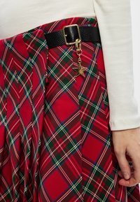 Rødt ternet nederdel med grønne og hvide linjer, et sort tekstureret bælte med en guldfarvet stjernevedhæng og plisseret stof for ekstra tekstur.