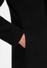 Abrigo negro con un tejido suave y texturizado, con un bolsillo lateral oculto. La manga presenta un detalle de puño limpio y entallado.