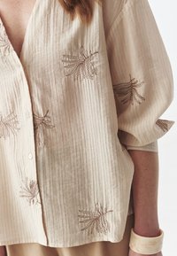 Camisa de botón color beige claro con rayas verticales, que presenta patrones florales bordados en marrón y mangas anchas y sueltas. Textura suave.