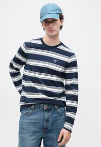 Tengerészkék és fehér csíkos hosszú ujjú póló kerek nyakkivágással, világoskék farmernadrággal és világoskék sapkával párosítva.