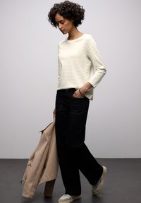 Hellcremefarbenes langärmliges Hemd mit Streifen, dunkelblaue Jeans mit weitem Bein, beiger Mantel über dem Arm, und helle Sneaker. Einfacher, lässiger Look.