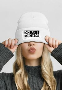 White knit beanie with a black patch reading "ICH HASSE MONTAGE" and a cat graphic; worn slightly pulled down, showing a gray sweater underneath.