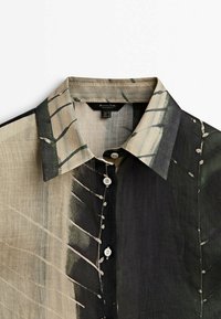 Chemise boutonnée avec un motif abstrait de coups de pinceau noirs, beiges et verts, avec un col pointu et des boutons blancs.
