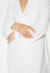 Vestido branco texturizado com um decote em V profundo, mangas longas com punhos franzidos e um acabamento geral de tecido suave.