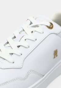 Sneaker in pelle bianca con lacci bianchi, occhielli dorati e piccolo logo dorato sul lato, su uno sfondo chiaro.