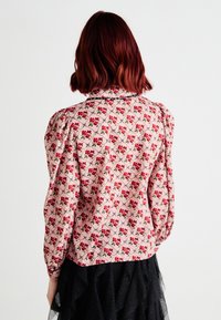 Blusa a maniche lunghe in tessuto rosa con un motivo floreale che presenta fiori rossi e verdi. Spalle a sbuffo e design con chiusura a bottoni sul retro.