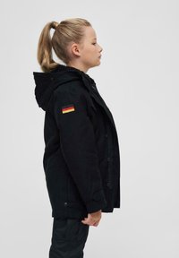 Črna parka jakna s kapuco, ovratnikom podloženim s flisom in stranskimi žepi. Na levi rokavu je majhen čin nemške zastave.