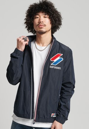 Giacca in nylon blu marino con polsini elastici, bordatura bianca della zip e un logo ricamato rosso e blu sul petto. Presenta tasche laterali.