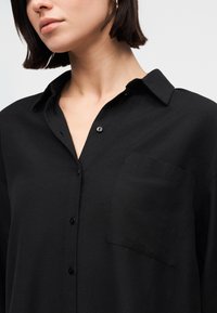 Žena oblečená v černé košili na knoflíky s náprsní kapsou a límcem, jejíž část krku a brady je viditelná, má krátké tmavé vlasy.