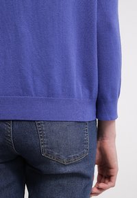 Fioletowy sweter z dzianiny o teksturowanym wykończeniu i ściągaczami na rękawach, w zestawieniu z ciemnoniebieskimi dżinsami z detalem na tylnej kieszeni.