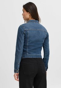 Chaqueta de mezclilla en azul oscuro, diseño corto con cuello clásico, mangas largas y cierre de botones en los puños. Pantalones negros con bolsillos traseros visibles.