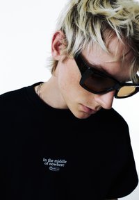 Mustat puuvillapaidat, joissa on valkoinen teksti "Keskellä ei mitään", yhdistettynä neliömäisiin mustiin aurinkolaseihin. Lyhyet vaaleat hiukset, pisamainen iho.