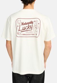 Cream-colored t-shirt with a reddish graphic on the back featuring the text "Naturally Lucky Club," dice, and crossed fingers design.